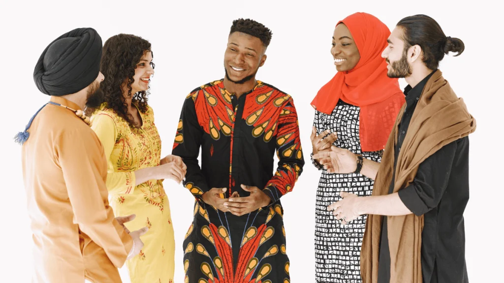 Grupo de cinco personas jóvenes de diferentes orígenes étnicos y culturales conversando animadamente en un entorno neutro y luminoso, todos vestidos con ropa tradicional representativa de sus culturas. Uno lleva turbante y kurta, otra viste un traje amarillo bordado, un hombre en el centro usa una túnica de colores rojo y negro, una mujer lleva hijab rojo y vestido estampado en blanco y negro, y otro hombre luce ropa oscura con una bufanda marrón sobre los hombros. La escena transmite armonía, respeto mutuo y colaboración intercultural, ilustrando visualmente el concepto de diplomacia cultural como herramienta para el entendimiento entre naciones y la construcción de puentes a través del diálogo y la identidad.