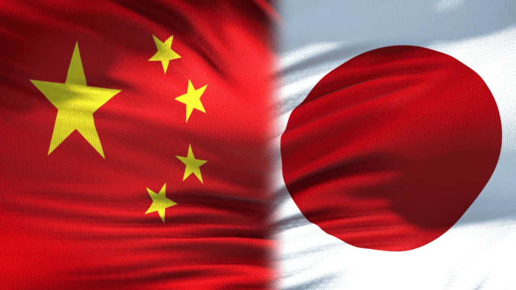 Primer plano de dos banderas ondeando unidas en el centro: a la izquierda, la bandera de China con fondo rojo y cinco estrellas amarillas en la esquina superior izquierda; a la derecha, la bandera de Japón con fondo blanco y un gran círculo rojo en el centro. Ambas telas muestran pliegues y textura, simbolizando la relación y vínculo entre ambos países asiáticos.
