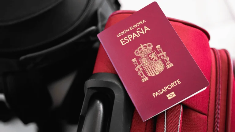 Primer plano de un pasaporte español de color burdeos, con el escudo nacional de España y las palabras “Unión Europea”, “España” y “Pasaporte” impresas en dorado en la portada. El pasaporte está apoyado sobre una maleta roja de tela con detalles en gris, junto a otra maleta negra parcialmente visible. La imagen transmite una temática de viaje o preparación para un desplazamiento internacional.