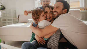 Una familia sonriente se abraza con cariño en una sala luminosa. La madre y el padre están sentados en el suelo mientras sostienen a sus dos hijos pequeños, uno en brazos y otro de pie detrás de ellos, todos con expresiones de alegría y afecto. La escena transmite calidez y unidad familiar, y puede representar el bienestar que conlleva obtener la residencia para familiares de españoles, permitiendo la convivencia cercana y legal en el país.